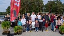 Grupa dorosłych i dzieci ustawiona razem na tle drzew i flagi z herbem, po bokach młode sadzonki drzew, w tle widoczni są inni uczestnicy oraz zabudowania rekreacyjne.