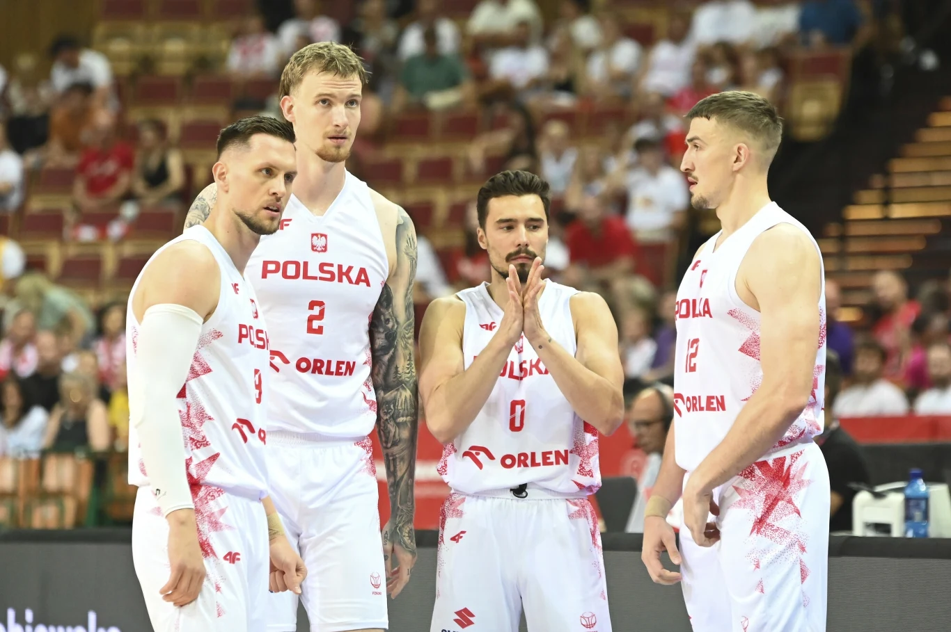 Czterech koszykarzy reprezentacji Polski w białych strojach z czerwonymi nadrukami stoi na boisku podczas meczu, za nimi widać zaciekawioną publiczność na trybunach.