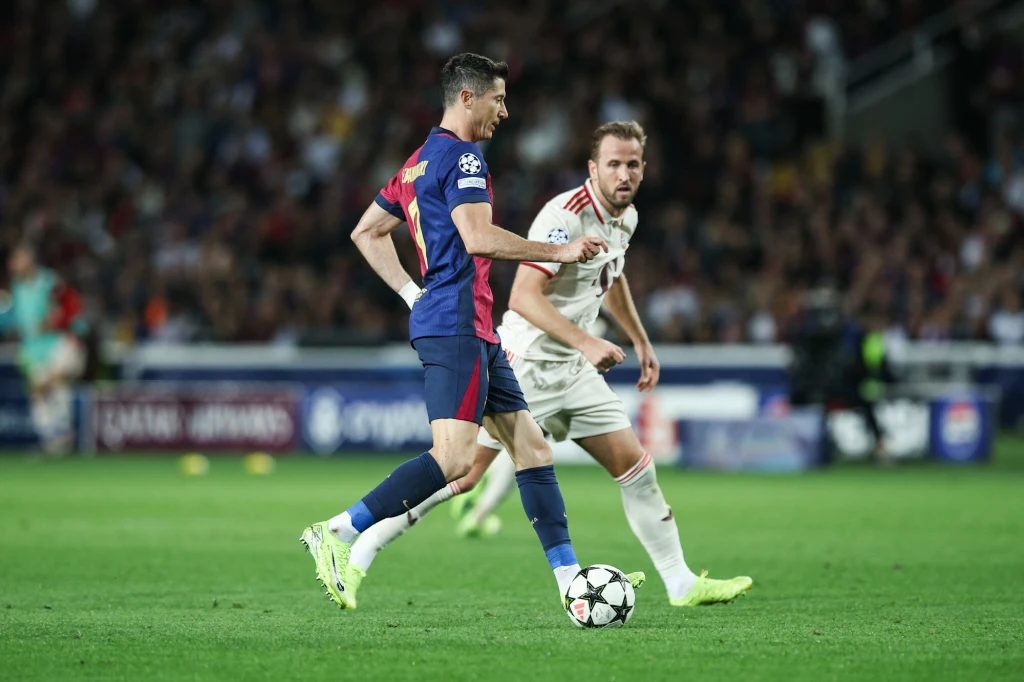 Robert Lewandowski i Harry Kane Dwóch piłkarzy w trakcie meczu piłkarskiego, jeden z zawodników prowadzi piłkę przy nodze, a drugi stara się go zatrzymać, wokół widać rozmyte postacie kibiców na trybunach oraz zieloną murawę stadionu.