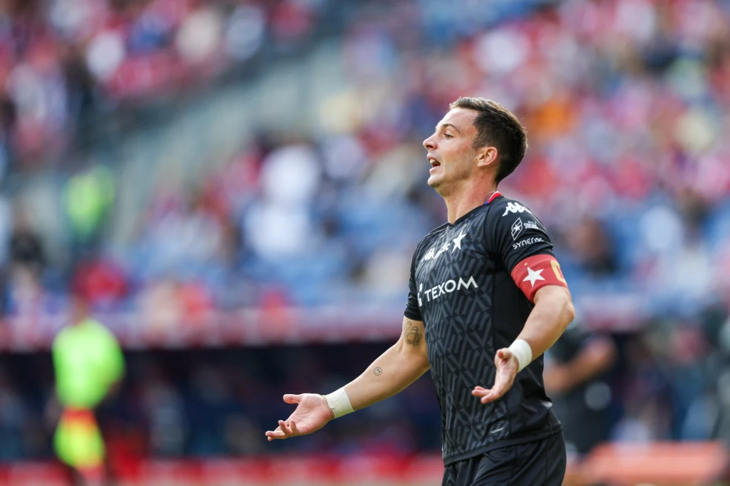 Triumfujący Angel Rodado (Wisła Kraków) w meczu ze Śląskiem Wrocław Piłkarz w czarnej koszulce i opasce kapitana na ramieniu z rozłożonymi rękami podczas meczu piłkarskiego na stadionie, w tle widoczni są rozmyci kibice.