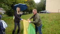 Dwie osoby wspólnie segregujące śmieci na trawniku, jedna trzyma parasol, podczas gdy druga pomaga rozkładać kolorowe worki na odpady, w tle widoczna zieleń i zaparkowane samochody.