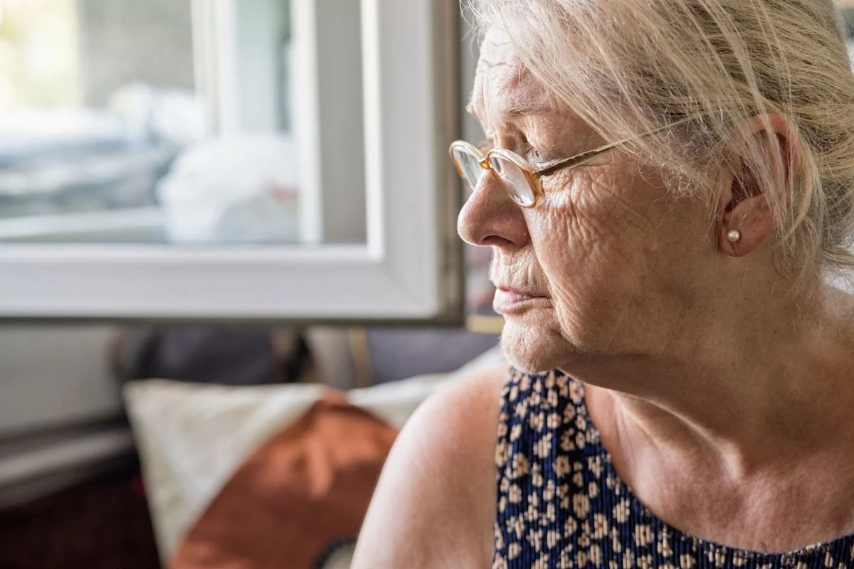 Starsza kobieta w okularach, siedząca w pomieszczeniu i spoglądająca przez okno, w tle widoczne rozmyte elementy wnętrza.