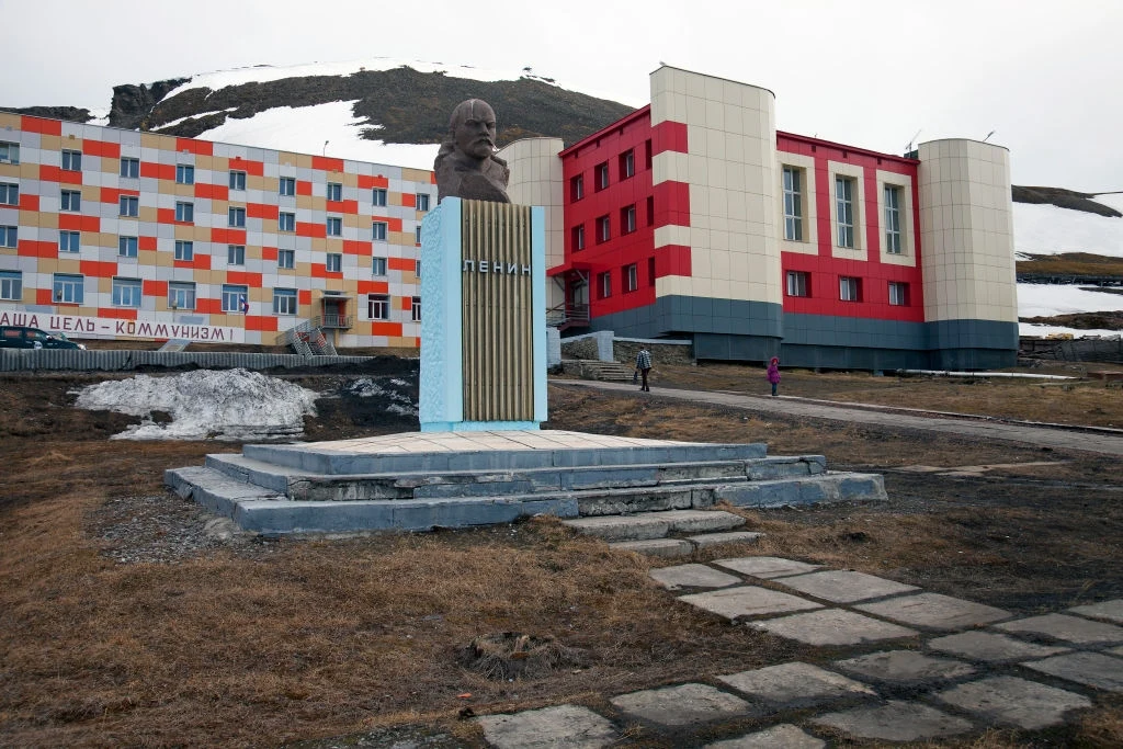 Pomnik Lenina w Barentsburgu Popiersie Lenina na tle kolorowego budynku z czerwonymi, białymi i pomarańczowymi elementami, z widocznymi pozostałościami śniegu na ziemi oraz górzystym krajobrazem w tle.