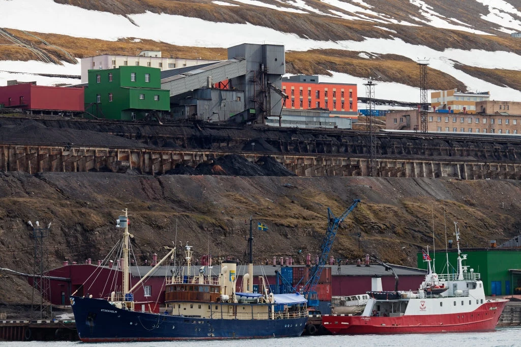 Kopalnia węgla w Barentsburgu Przemysłowy port z dwiema zacumowanymi łodziami u brzegu, w tle charakterystyczne kolorowe budynki przemysłowe oraz zaśnieżone wzgórza.