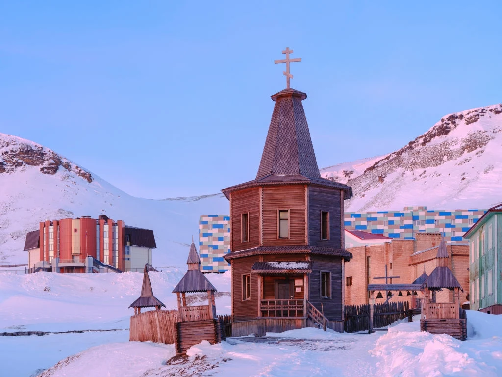Cerkiew w Barentsburgu Drewniana cerkiew z krzyżem na dachu stoi na śnieżnym tle, otoczona płotem, w sąsiedztwie kolorowych budynków i górskiego krajobrazu.