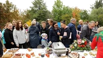 Stół z białym obrusem uginający się od różnorodnych potraw, ciast i przekąsek, dookoła duża grupa ludzi w różnym wieku ustawiająca się w kolejce, w tle drzewa i zieleń, po prawej stronie kompozycja kwiatowa.