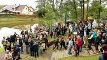 Tłum ludzi zgromadzony w parku nad małym stawem bierze udział w sadzeniu drzewa, uczestnicy stoją zarówno na trawie, jak i na brukowanym chodniku, wokół panuje atmosfera zaangażowania i wspólnoty.