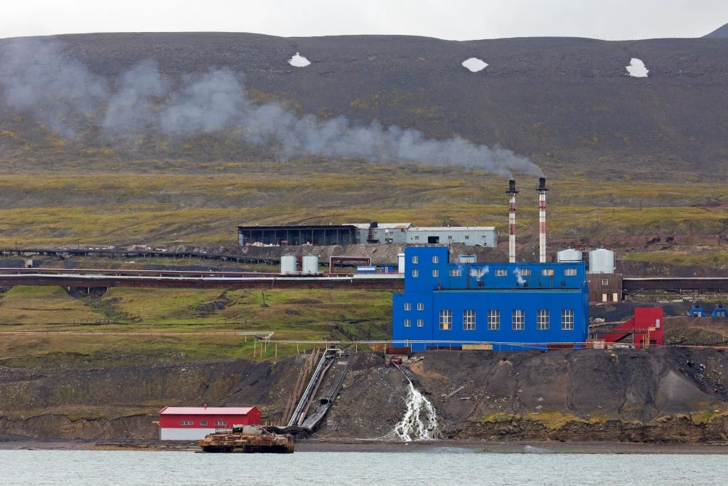 Elektrownia w Barentsburgu Obiekt przemysłowy z niebieskim budynkiem i dwoma kominami emitującymi dym, położony nad brzegiem wody, w tle pagórki pokryte niską roślinnością i resztkami śniegu.