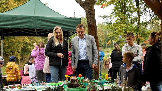 Grupa ludzi oglądająca prace plastyczne i makiety ustawione na stole podczas plenerowego wydarzenia pod zielonym namiotem, w otoczeniu jesiennych drzew, uczestnicy ubrani w kurtki wskazują na chłodniejszą pogodę, atmosfera przyjazna i rodzinna