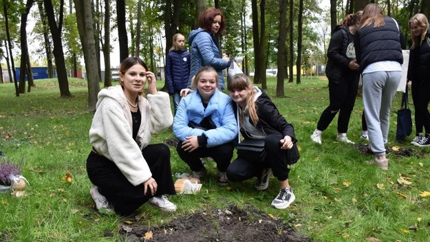 Grupa młodych osób zgromadzona w parku podczas wspólnej aktywności, trzy dziewczyny klęczą przy wykopanym dołku na trawie, w tle widać innych uczestników wydarzenia w jesiennym otoczeniu drzew.