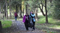 Dwie osoby w kolorowych kurtkach niosą czarny worek na śmieci, spacerując alejką w parku otoczonym drzewami, w tle widać kolejną osobę oddalającą się w stronę drzew.