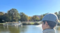 Chłopiec w czapce i kamizelce wędkarskiej z napisem 'GRUPY WĘDKARSKIE Catch & Release' stoi nad brzegiem jeziora z wędką w ręku, w tle rozpościera się spokojny krajobraz wodny i drzewa w jesiennej kolorystyce.