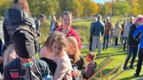 Dzieci wybierające kolorowe zabawki zrobione z plecionych sznurków przy stoliku ustawionym na zewnątrz, obok nich kobieta oraz grupa osób w tle na świeżym powietrzu.