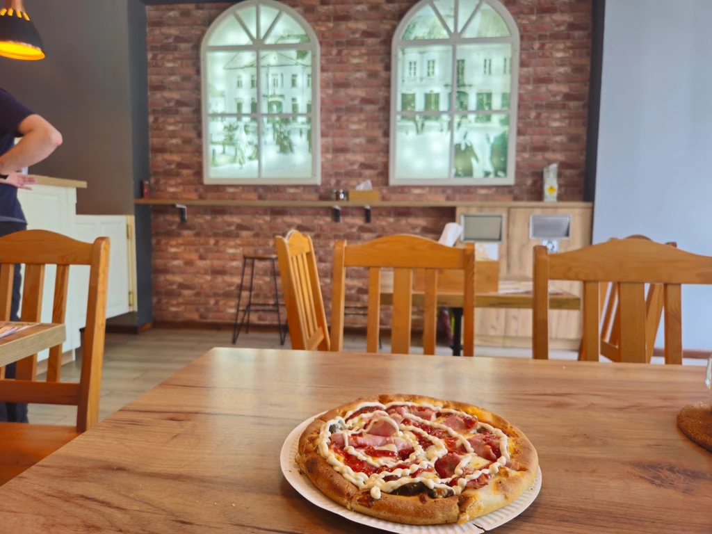 Pizza na talerzu ustawionym na drewnianym stole w przytulnej restauracji z drewnianymi krzesłami, ceglanym murem oraz dużymi oknami z widokiem na ulicę i klasyczną zabudowę.