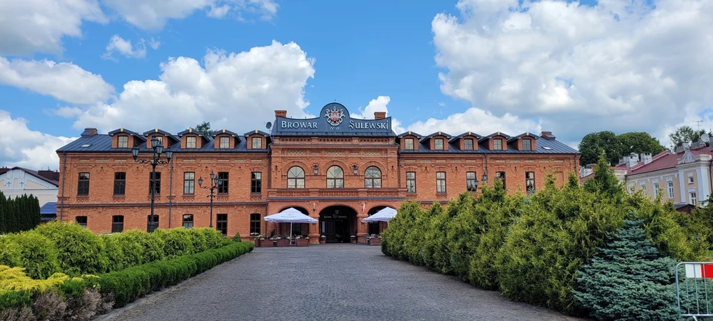 Ceglany budynek browaru z ozdobnym napisem na fasadzie, na pierwszym planie zadbany brukowany chodnik otoczony zielonymi krzewami, nad całością rozciąga się błękitne niebo z białymi chmurami.