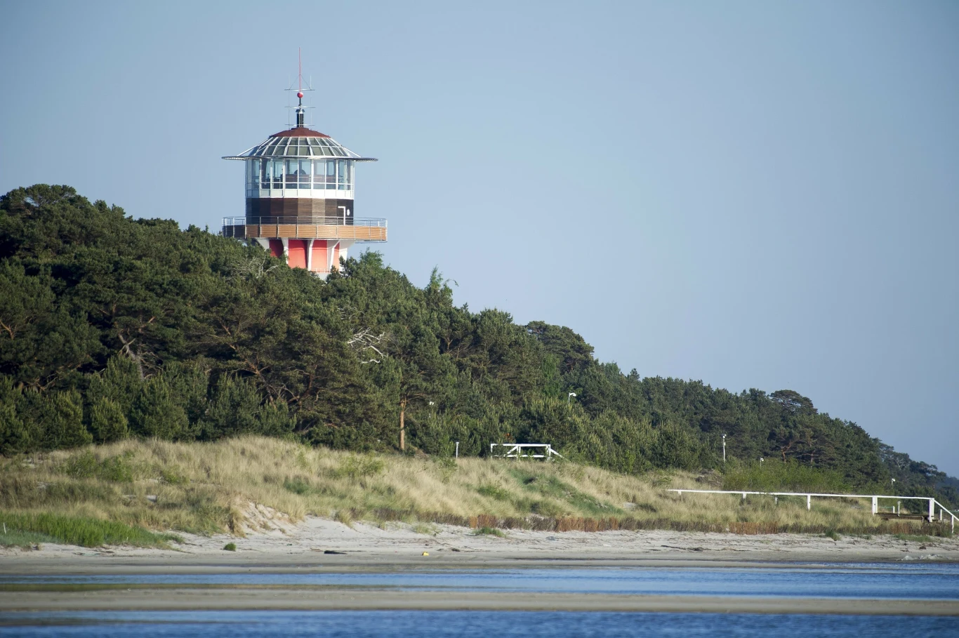 Rezydencja Prezydenta RP na Mierzei Helskiej Latarnia morska położona nad linią lasu, otoczona zielonymi drzewami, w tle jasnoniebieskie niebo oraz piaszczysta plaża na pierwszym planie.