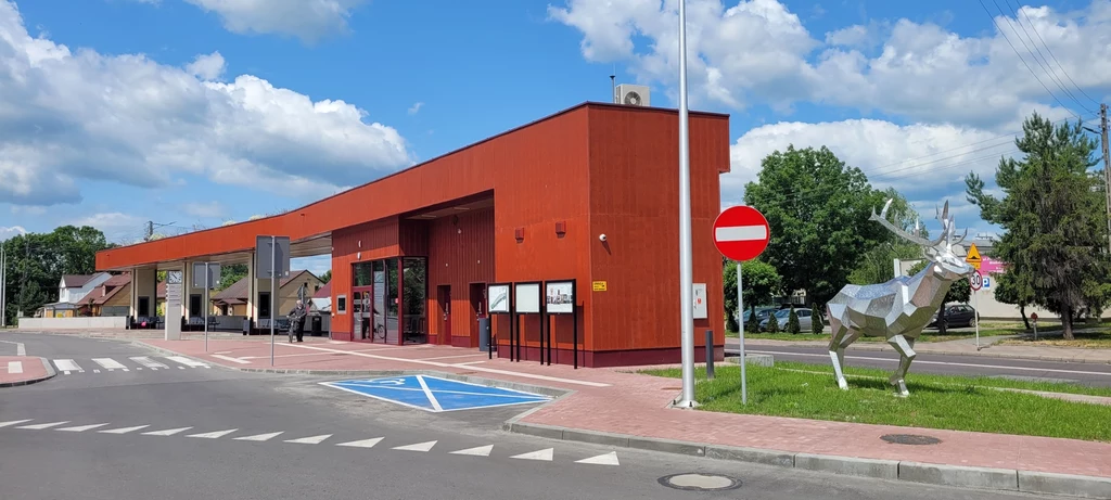Nowoczesny, czerwony budynek stacji autobusowej na tle nieba z białymi chmurami, przy którym znajduje się metalowa rzeźba jelenia z różowym nosem oraz znak drogowy zakaz wjazdu. W pobliżu widoczny jest parking z wyznaczonym miejscem dla osób niepełnosp...