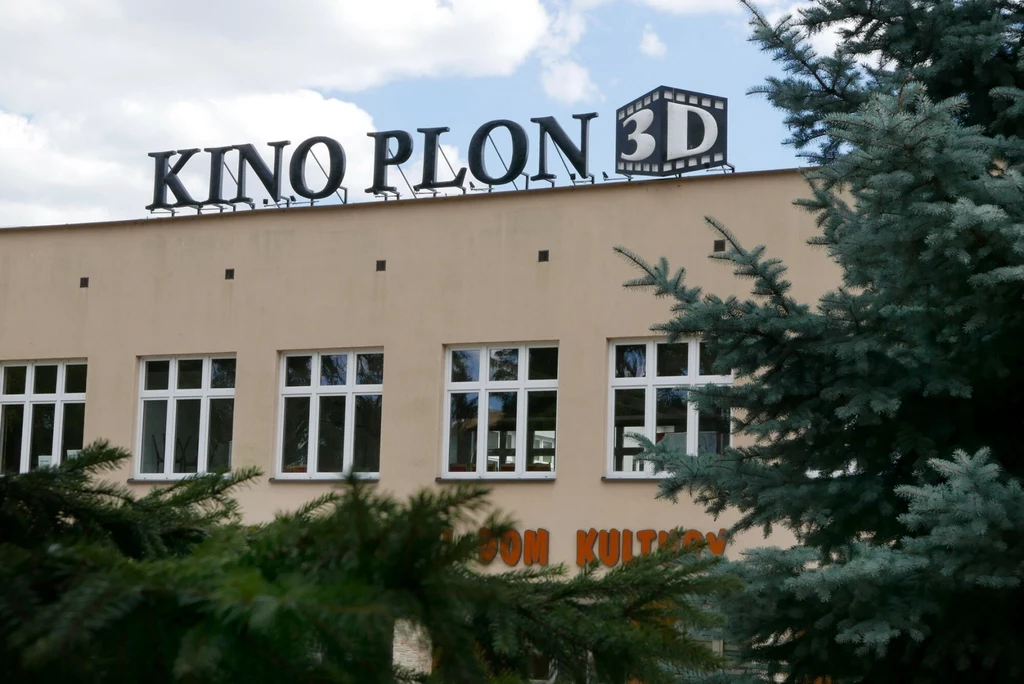 Jasny budynek z widocznym napisem KINO PLON 3D oraz częściowym napisem DOM KULTURY, otoczony zielonymi iglakami na pierwszym planie; klarowne niebo z chmurami w tle.