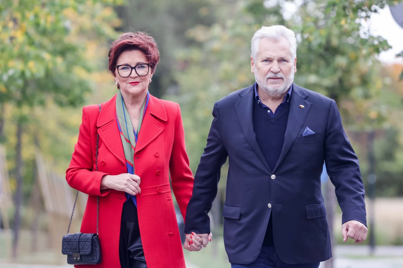 Jolanta i Aleksander Kwaśniewscy Elegancka kobieta w czerwonym płaszczu i mężczyzna w granatowej marynarce idą razem, trzymając się za ręce, na tle zieleni i drzew.
