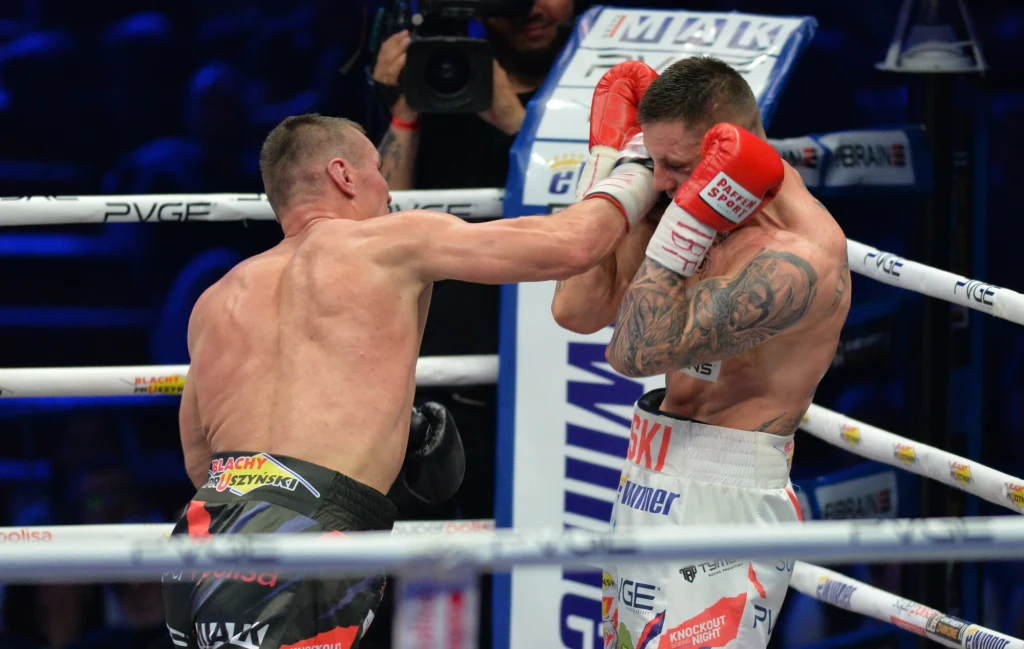 Dwóch bokserów podczas intensywnej walki w ringu, jeden z zawodników zadaje cios, podczas gdy drugi zasłania twarz rękawicami. W tle widoczny sędzia i publiczność. Najważniejsze są dynamiczne emocje oraz napięcie pojedynku.