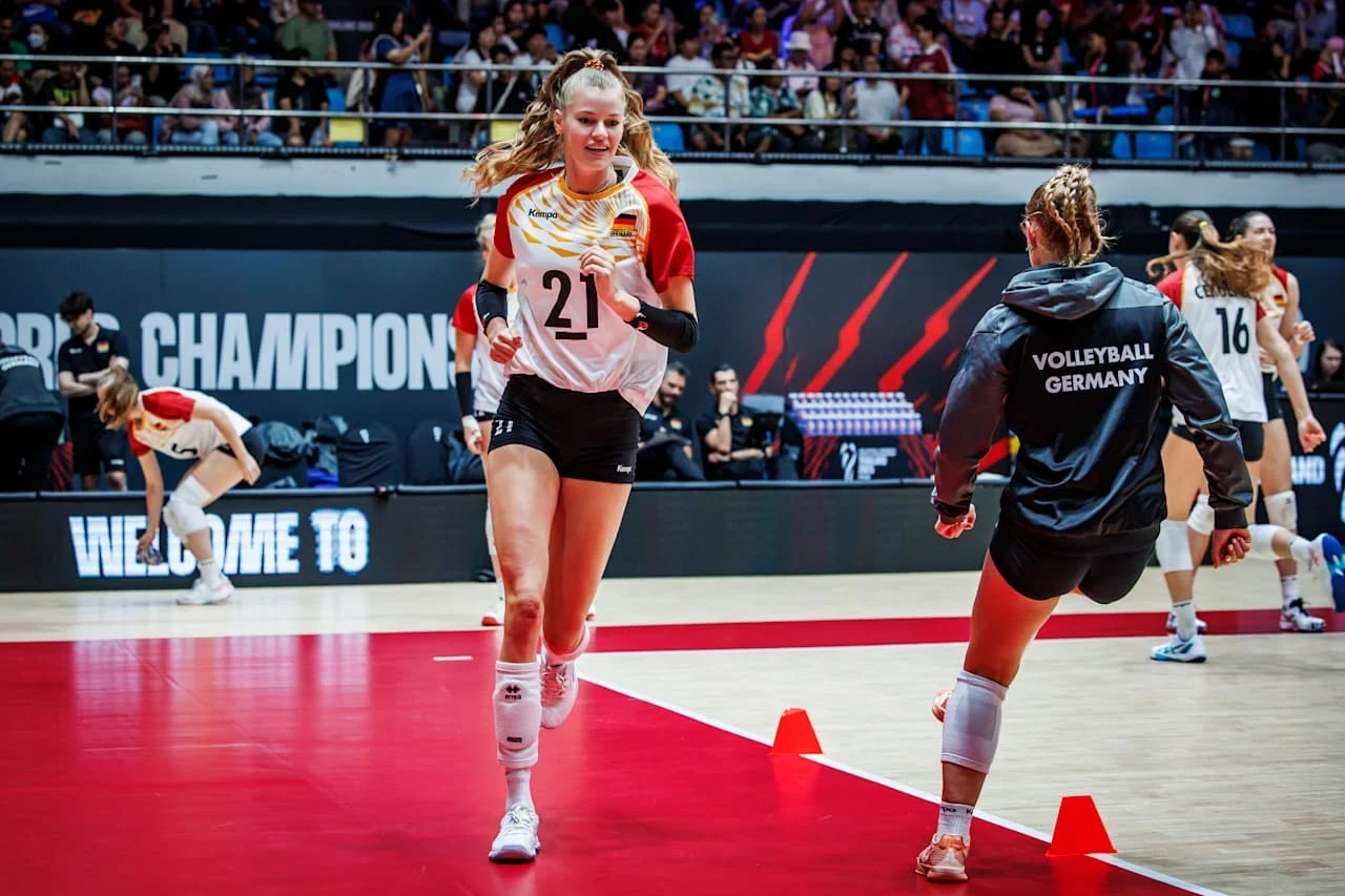 Siatkarki reprezentacji Niemiec Zawodniczka drużyny siatkarskiej Niemiec w trakcie rozgrzewki na hali sportowej, na pierwszym planie widoczna jest energiczna interakcja z inną zawodniczką, w tle widać publiczność i napis 'WELCOME TO' na elektronicznym banerze.