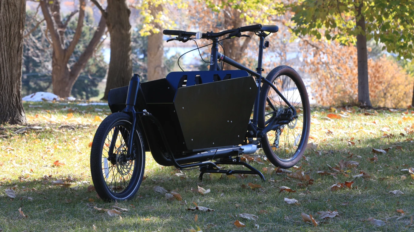 Istnieje sposób, aby zwykły rower przerobić na rower cargo Czarny rower transportowy z dużą skrzynią ładunkową umieszczoną z przodu stoi na trawie w parku jesienią, w otoczeniu drzew o kolorowych liściach.