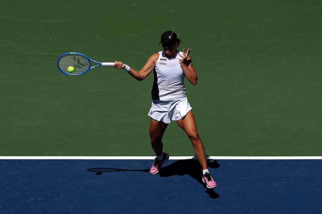 Iga Świątek, US Open 2025 Tenisistka w białym stroju uderza piłkę na zielono-niebieskim korcie, skupiona na precyzyjnym wykonaniu zagrania.