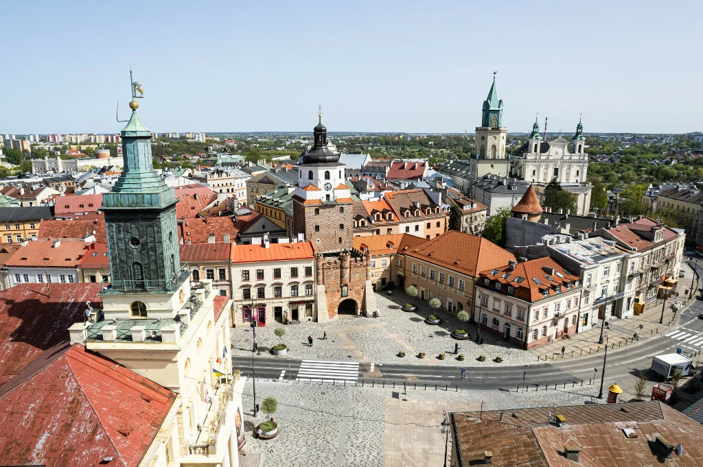 Panorama Starego Miasta w Lublinie, widoczne charakterystyczne kamienice, Brama Krakowska oraz wieże kościołów otoczone czerwonymi dachami, w tle współczesna zabudowa miejska.
