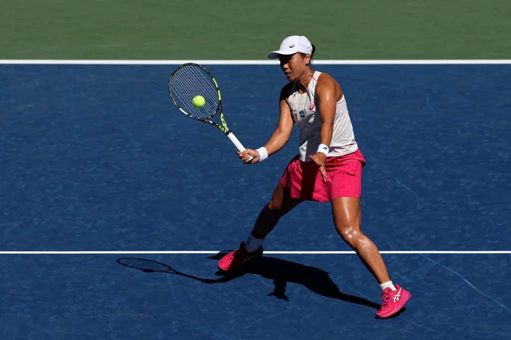 Janice Tjen Tenisistka w sportowym stroju podczas rozgrywki, wykonuje uderzenie rakietą w kierunku nadlatującej piłki tenisowej na niebieskim korcie.