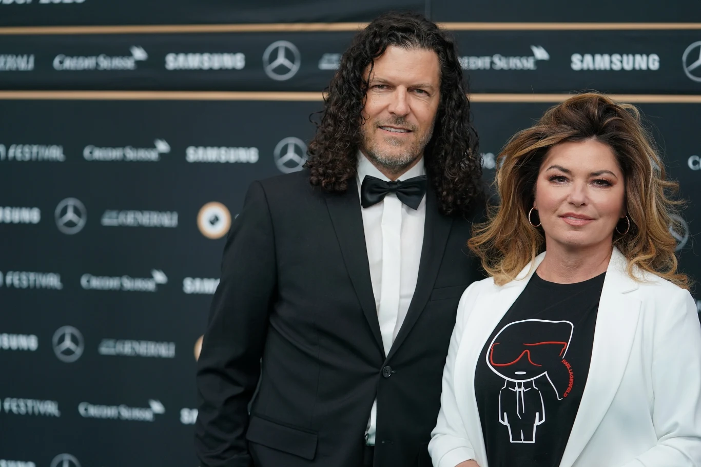 Shania Twain i Frederic Thiebaud w 2020 r. Dwie elegancko ubrane osoby pozujące razem na tle ścianki promocyjnej z logotypami znanych marek, mężczyzna w smokingu i kobieta w białej marynarce oraz koszulce z charakterystycznym nadrukiem.