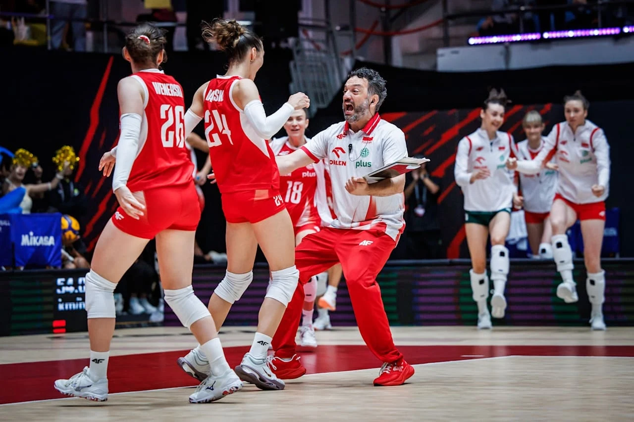 Trener w biało-czerwonym dresie energetycznie prowadzi drużynę siatkarek podczas meczu, zawodniczki w czerwonych i białych strojach komunikują się między sobą na środku boiska, w tle widać kolejne zawodniczki wchodzące na parkiet.