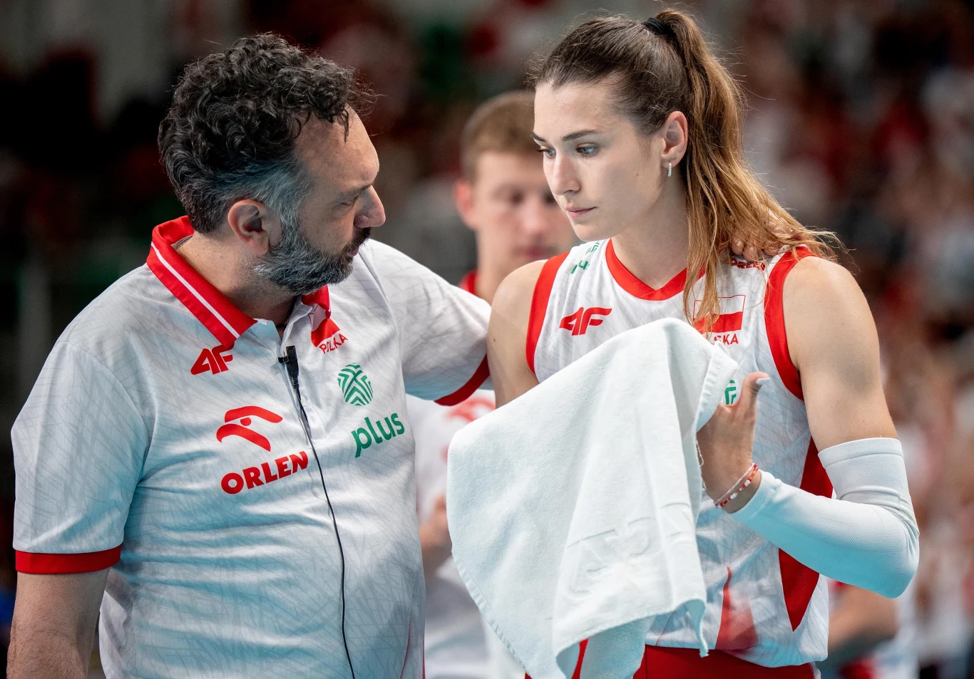 Trener rozmawia z siatkarką w biało-czerwonym stroju, która trzyma ręcznik, oboje wyglądają na skoncentrowanych, w tle niewyraźna hala sportowa i publiczność.