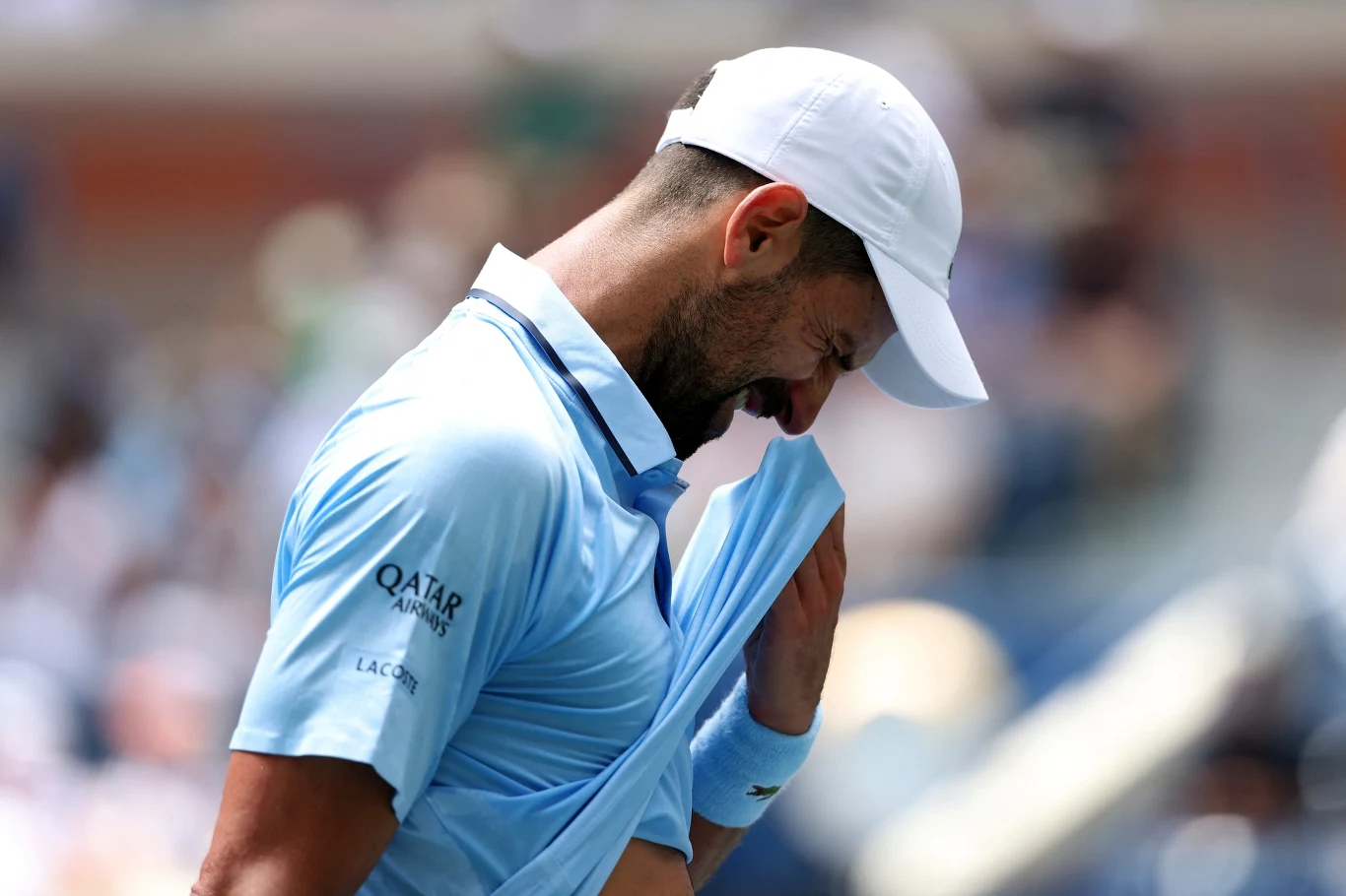 Sportowiec ubrany w jasnoniebieską koszulkę i białą czapkę z daszkiem trzyma koszulkę przy twarzy w wyrazie zmęczenia lub emocji, tło rozmyte, skupienie na postaci.