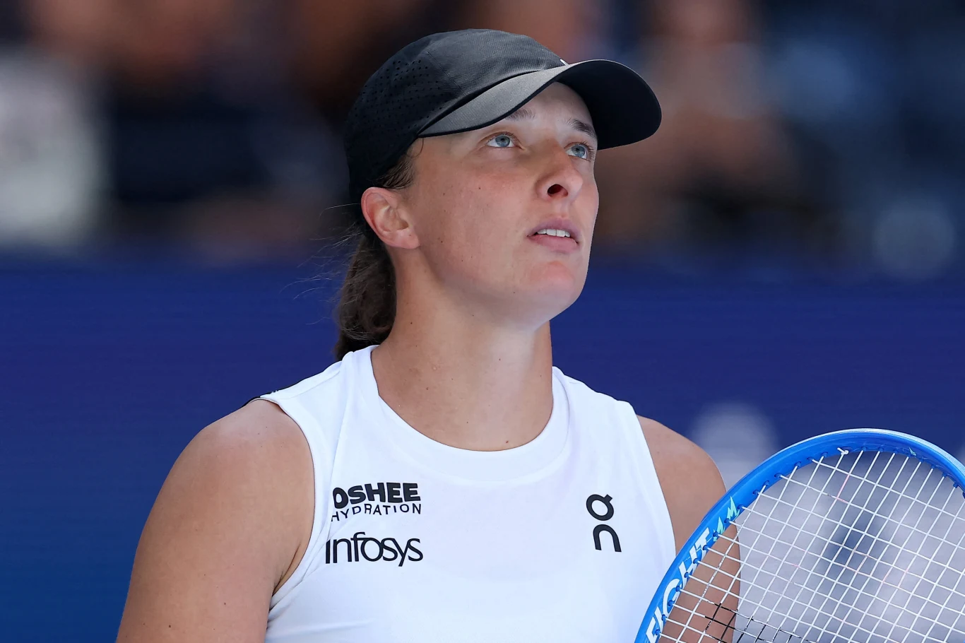 Tenisistka w białym stroju sportowym i czarnej czapce z daszkiem, trzymająca rakietę tenisową i patrząca w górę, na tle rozmazanej, niebieskiej areny sportowej.