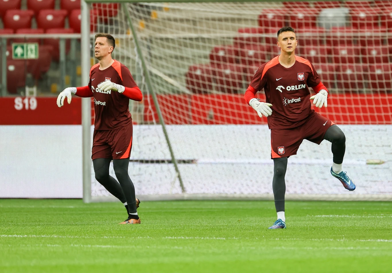Wojciech Szczęsny i Mateusz Kochalski podczas treningu reprezentacji Polski Dwóch piłkarzy w czerwonych strojach treningowych wykonuje ćwiczenia na murawie stadionu piłkarskiego, w tle widoczne czerwone krzesełka trybun oraz siatka bramki.