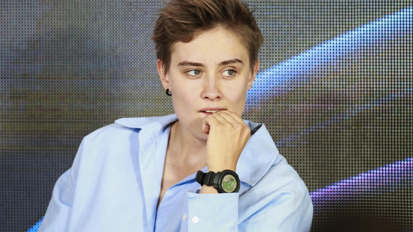 Młoda osoba z krótkimi włosami w jasnoniebieskiej koszuli, z zamyślonym wyrazem twarzy, opiera dłoń o usta i spogląda w bok, w tle rozmyte wzory świetlne.
