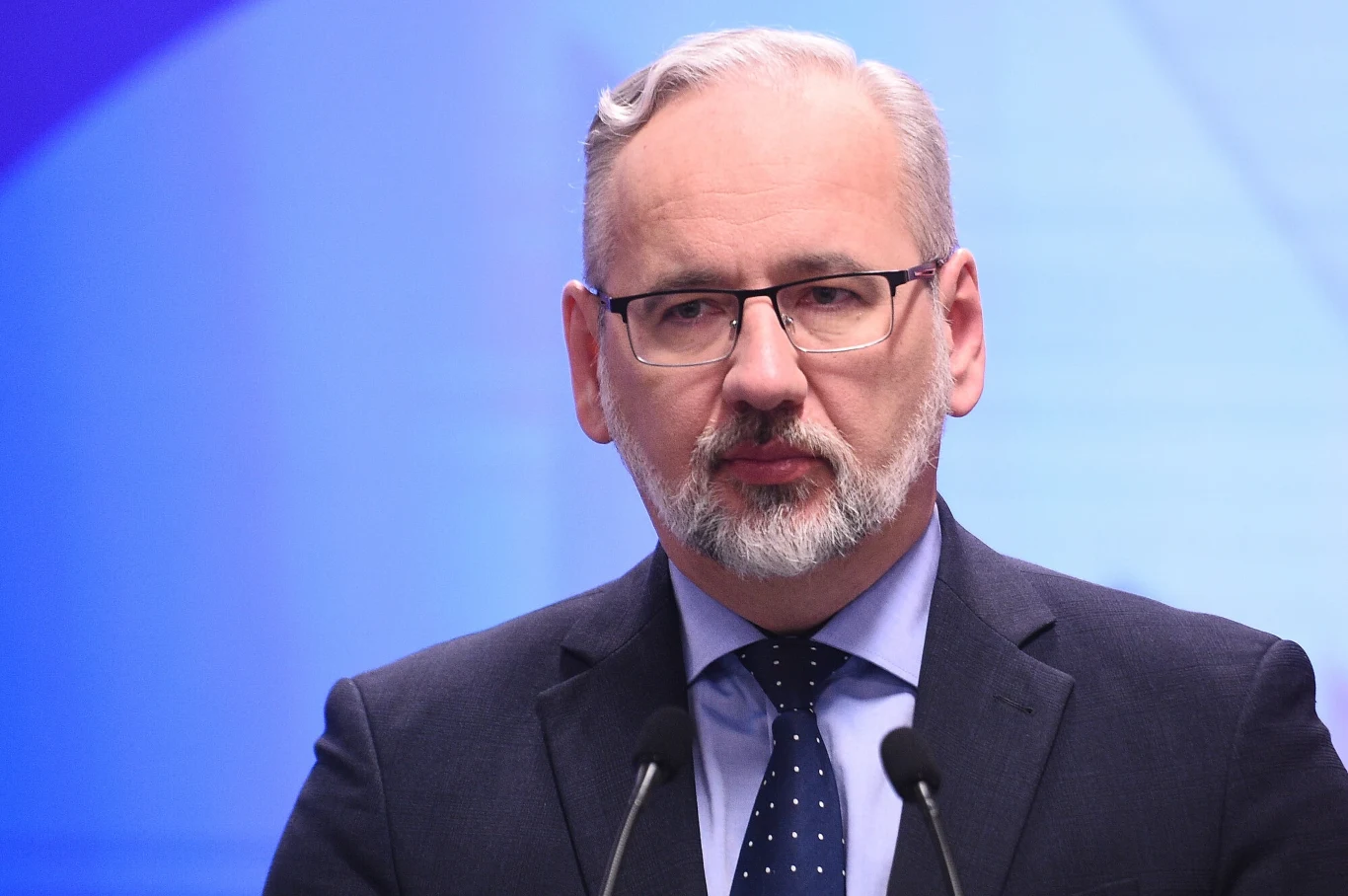 Adam Niedzielski zabrał głos po ataku Mężczyzna w garniturze i okularach stoi przed mikrofonem na tle niebieskiego ekranu, krótkie siwe włosy i broda, wyraz twarzy poważny.