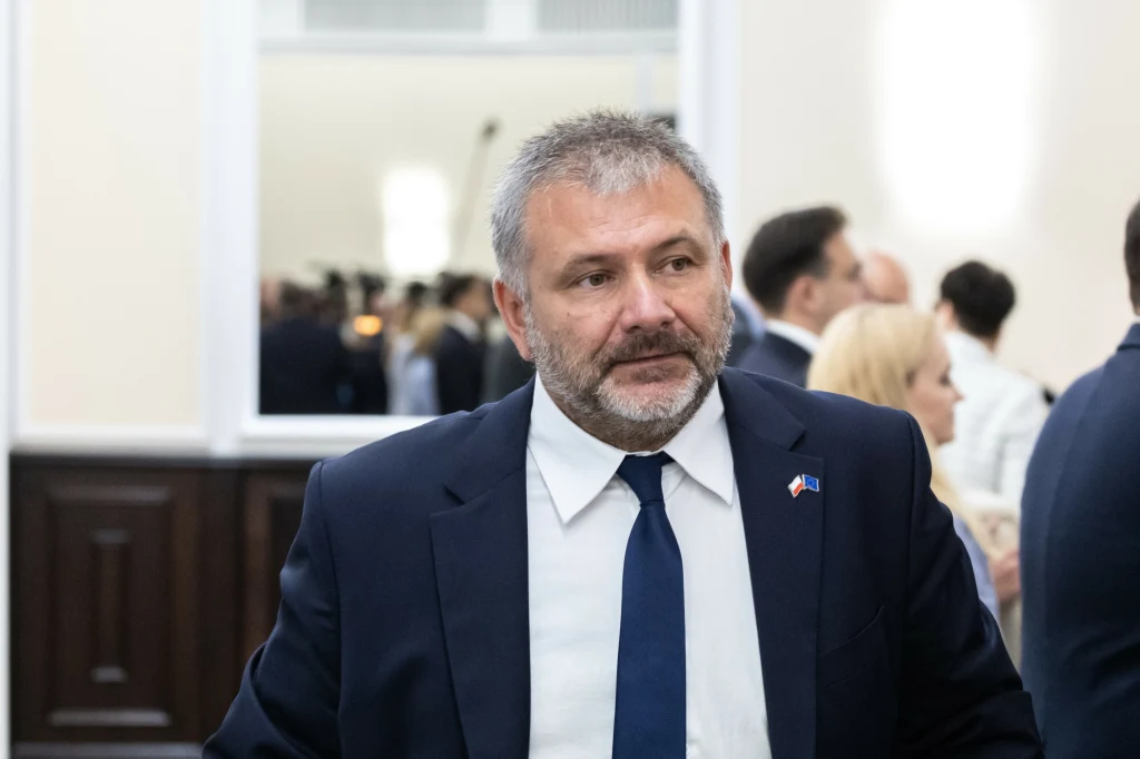 Szef MS Waldemar Żurek Mężczyzna w średnim wieku w garniturze i krawacie stoi w jasnym pomieszczeniu podczas oficjalnego spotkania lub konferencji, na drugim planie widoczne są sylwetki innych osób w eleganckim stroju.