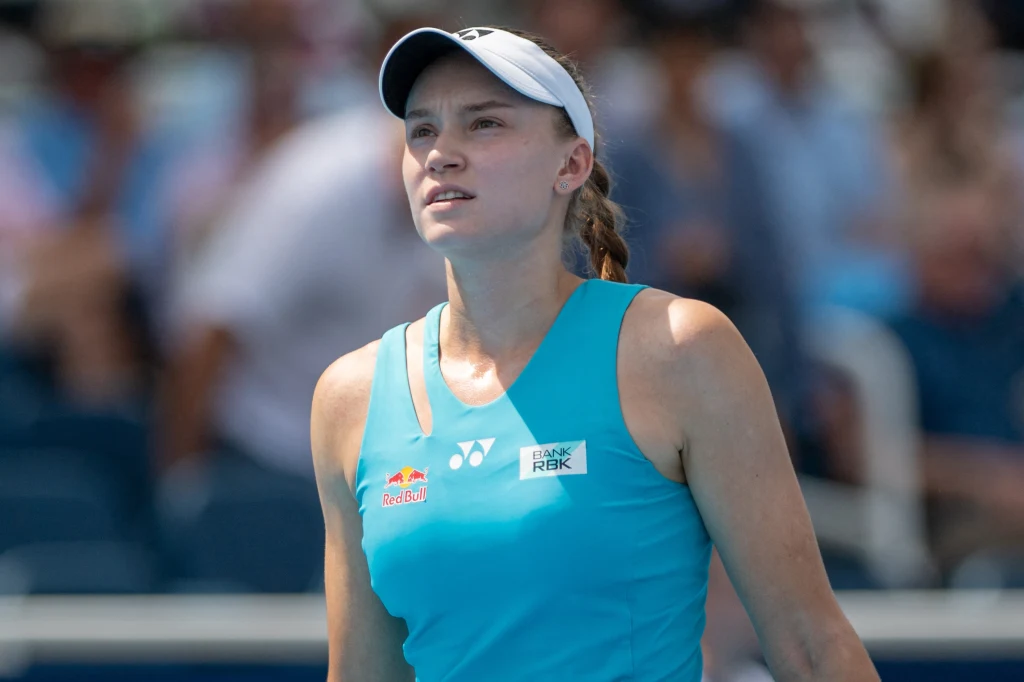 Młoda kobieta w sportowym stroju tenisowym z jasnoniebieską koszulką i białym daszkiem stoi na korcie tenisowym, skupiona i skoncentrowana, w tle widoczna rozmyta publiczność.