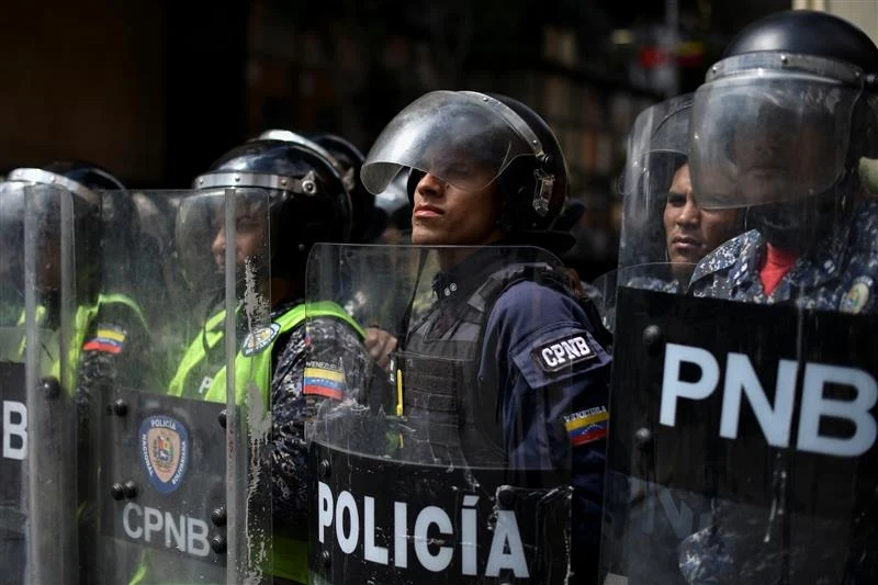 Wenezuela przygotowuje się na "agresję ze strony USA" Grupa funkcjonariuszy policji w strojach ochronnych oraz kaskach, trzymających przezroczyste tarcze z napisem 'POLICÍA' i 'PNB', skupionych w szeregu podczas służby. Na mundurach widoczne są naszywki i flagi Wenezueli.