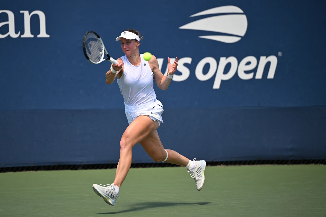 Zawodniczka ubrana w biały strój tenisowy wykonuje dynamiczne uderzenie rakietą podczas meczu na kortach turnieju US Open, w tle charakterystyczne logo turnieju i niebieska nawierzchnia kortu.