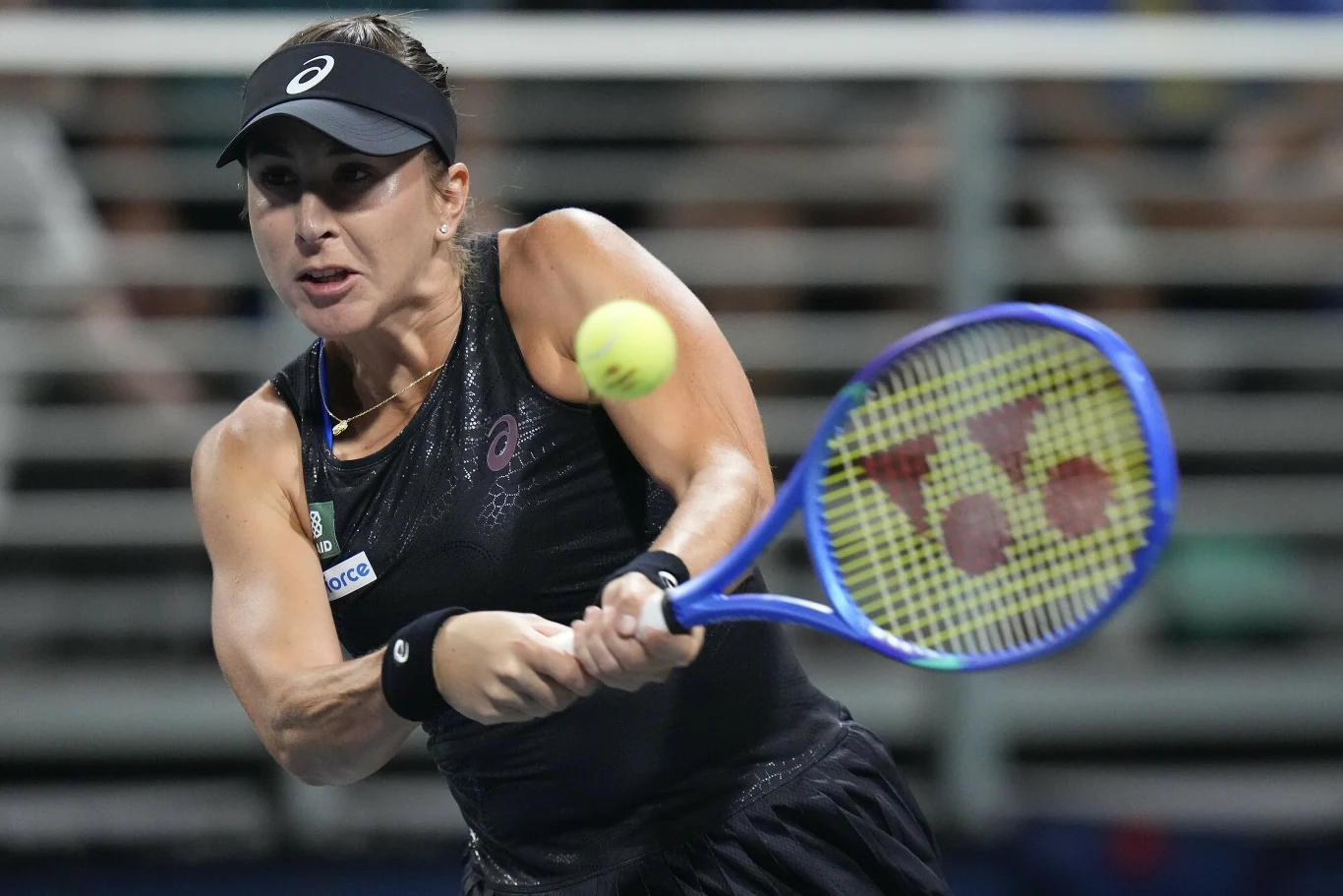Zawodniczka w czarnej sportowej odzieży odbija piłkę tenisową podczas meczu, trzymając rakietę tuż przed sobą, skoncentrowana na uderzeniu, w tle metalowe barierki kortu.