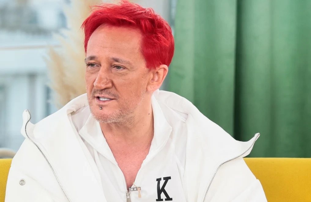 Michał Wiśniewski otwarcie mówi o samoakceptacji Mężczyzna z krótkimi, intensywnie czerwonymi włosami ubrany w biały strój z literą K na piersi, siedzi na żółtej sofie na tle zielonych zasłon.