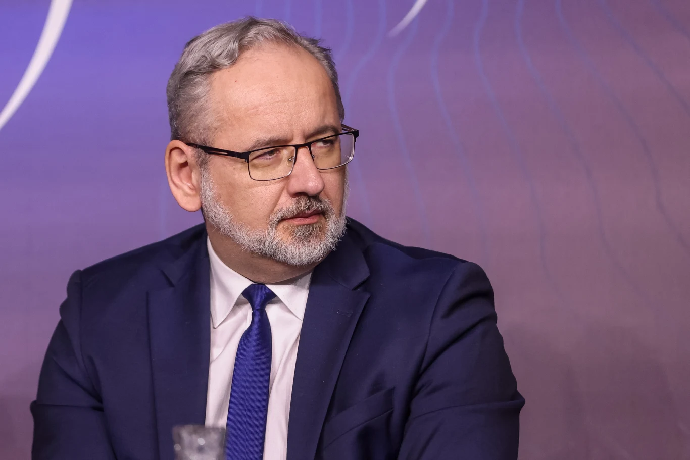 Nowe informacje policji ws. pobicia byłego ministra zdrowia Adama Niedzielskiego Mężczyzna w średnim wieku z siwymi włosami i brodą, ubrany w granatowy garnitur, białą koszulę i niebieski krawat, siedzi w pomieszczeniu o fioletowym tle, patrząc w bok.