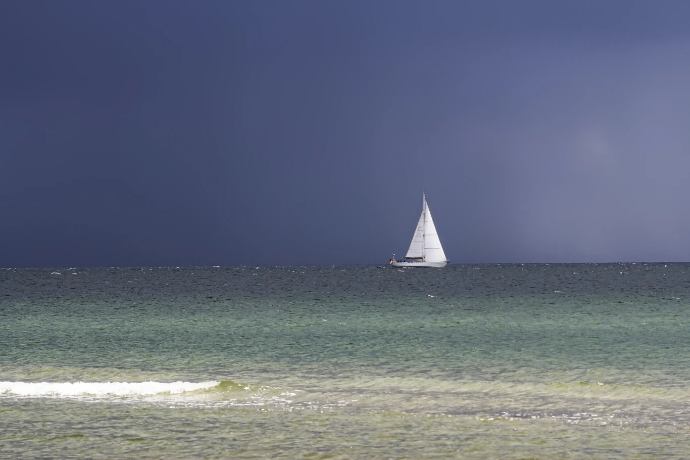 Jak głębokie jest Morze Bałtyckie i w którym miejscu znajdują się jego najgłębsze punkty? Zdj. ilustracyjne. Biała żaglówka płynie po spokojnym morzu pod ciemnym, burzowym niebem, kontrastując jasno na tle granatowej linii horyzontu