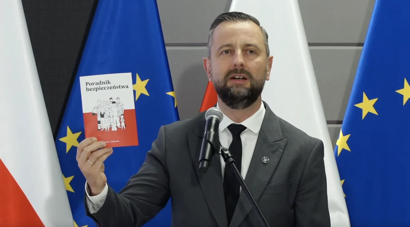 Poradnik bezpieczeństwa. Rząd prezentuje przewodnik na sytuacje kryzysowe. Mężczyzna w garniturze stojący przy mikrofonie trzyma w dłoni broszurę o tytule 'Poradnik bezpieczeństwa', w tle widoczne flagi Polski oraz Unii Europejskiej.