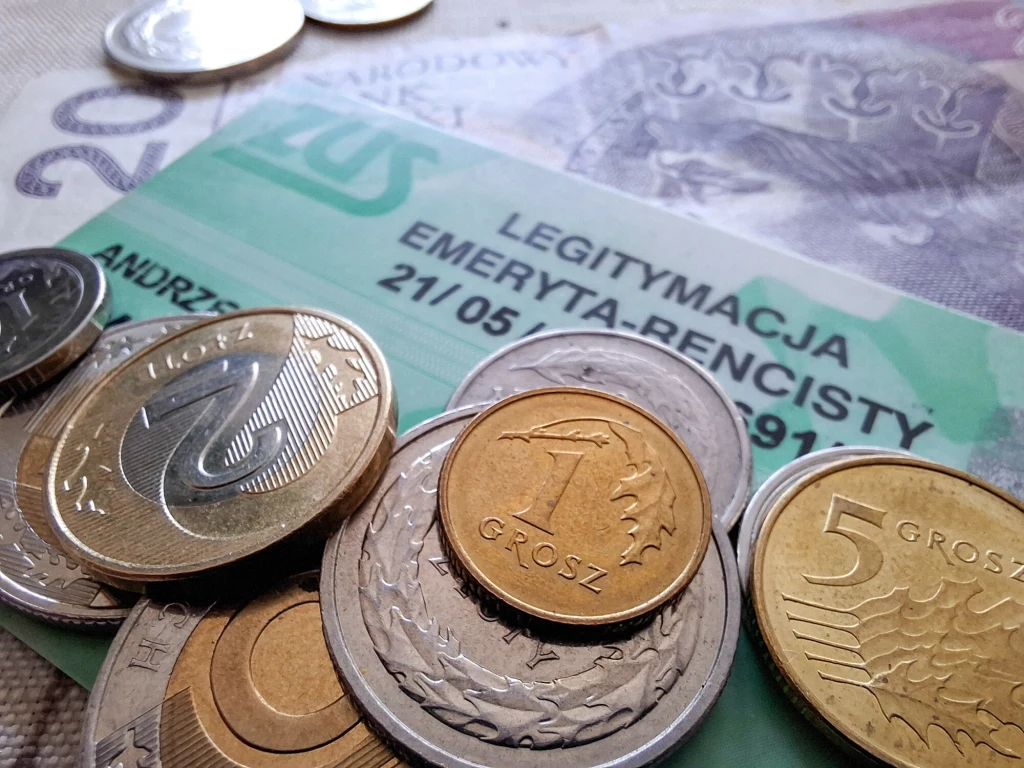 Polskie monety i banknoty leżą na zielonej legitymacji emeryta-rencisty ZUS, skupiając uwagę na kwestiach finansowych emerytów.