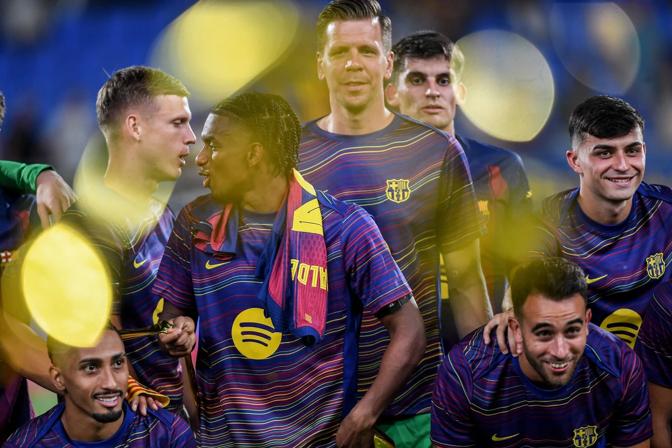 Grupa piłkarzy w kolorowych koszulkach sportowych z logo dużego sponsora, stojących razem na murawie, część z nich uśmiechnięta, wokół rozproszone żółte światła, nadające zdjęciu dynamiczny charakter.