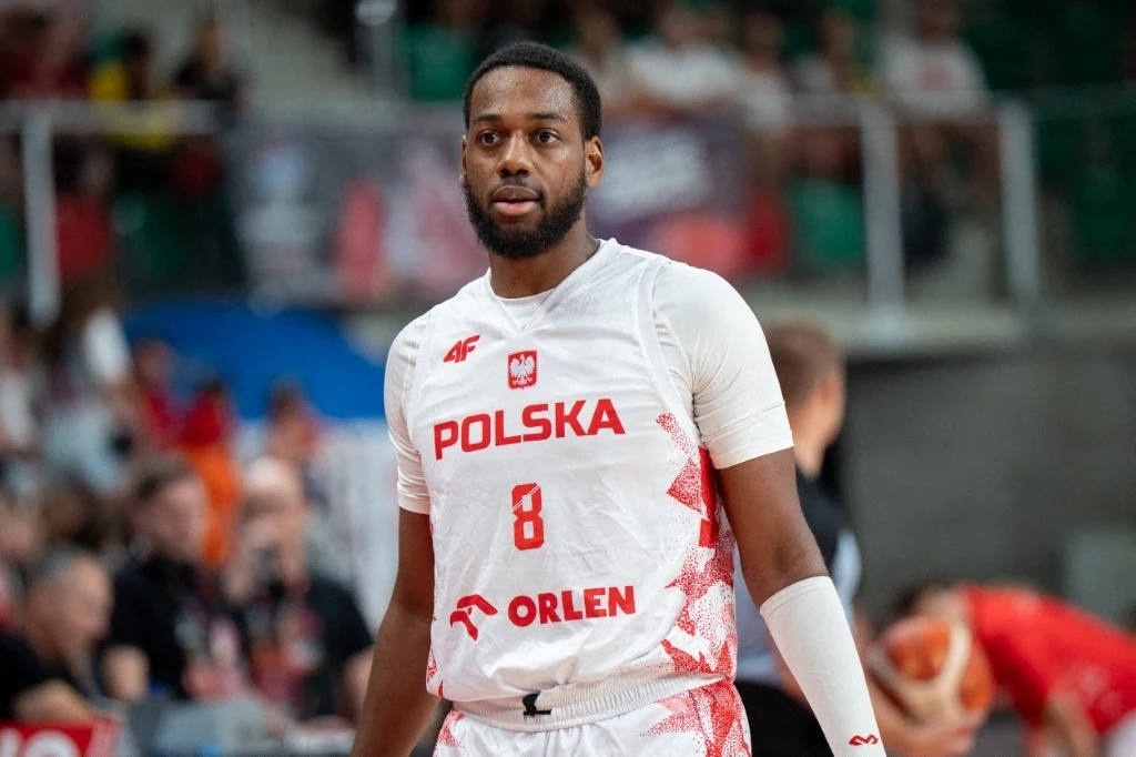 Koszykarz w białym stroju reprezentacji Polski z numerem 8 podczas meczu, w tle rozmyty tłum kibiców oraz hala sportowa.