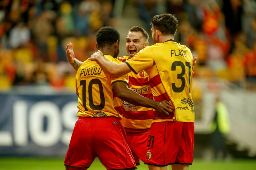 Jagiellonia Białystok Trzech piłkarzy w żółto-czerwonych strojach drużyny świętuje zdobycie gola na murawie stadionu, obejmując się i okazując radość, w tle widać rozmyte sylwetki innych kibiców i elementy trybun.